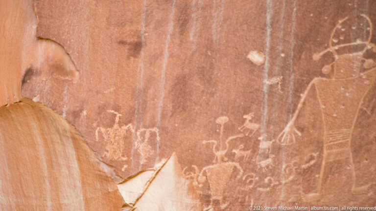 Pale human-like and geometric forms appear on red sandstone above a sloping rock ledge.
