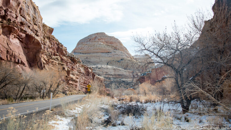 A paved road bends toward a pale dome between red cliffs and snow-dusted slopes.