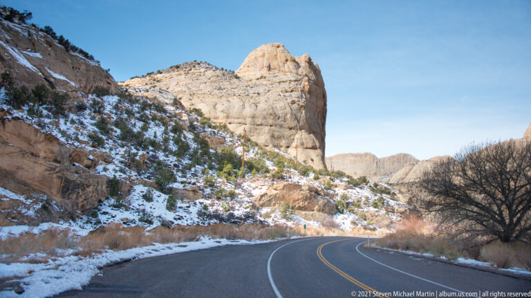 A double-yellow road curves below a pale sandstone dome and snow-dusted cliffs.