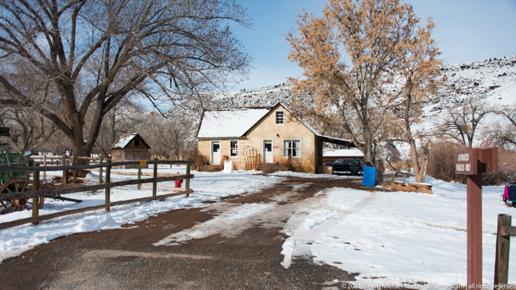A farmhouse and outbuildings stand among snowy orchard trees, fences, and a wooden wagon.