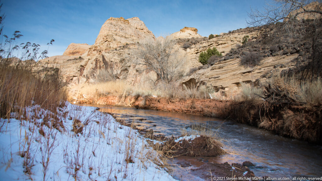 A shallow stream runs beside snow, dry grasses, and red sandstone banks.