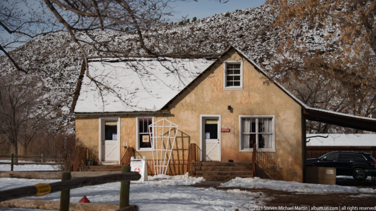 A tan building with a steep snowy roof, white-trimmed doors, and an OPEN sign stands behind a fence.