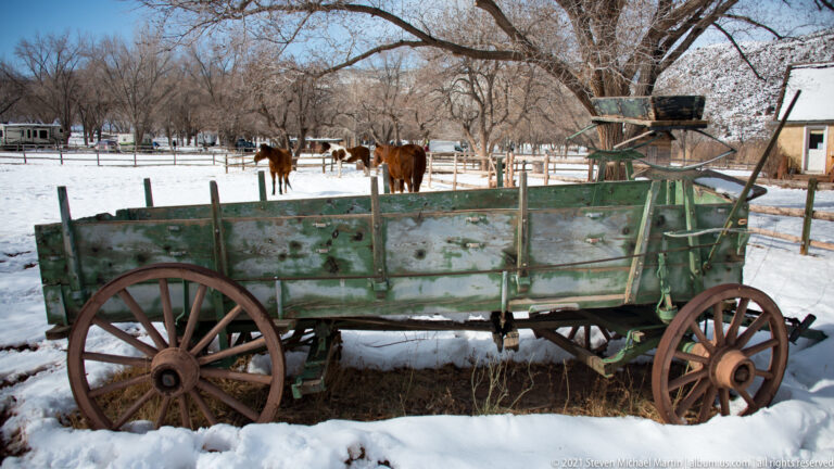 A green wooden-spoked wagon stands before fenced horses in a snowy orchard.