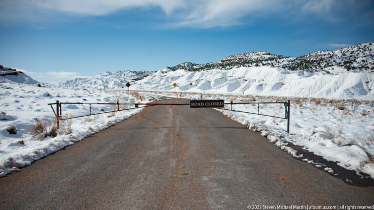 A ROAD CLOSED barrier blocks a paved road bordered by snow-covered slopes.