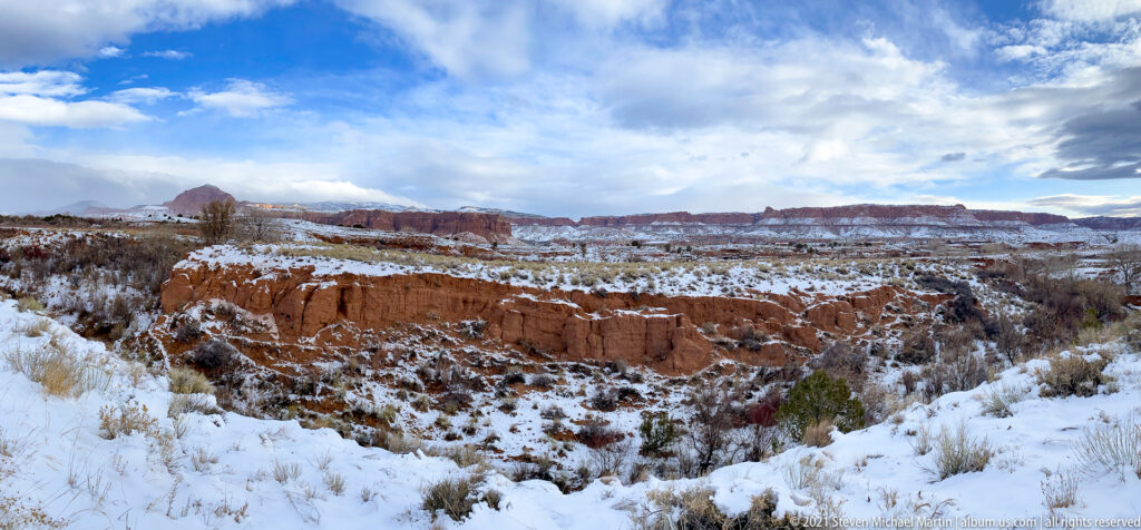 Snow covers red-rock benches and gullied badlands, with sparse shrubs below a broad cloudy sky.