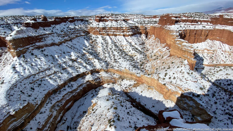 A broad canyon bend curves around a snowy basin below a sunlit red cliff.