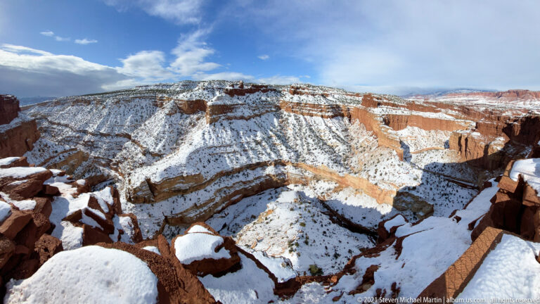 Snow-covered sandstone slabs frame a steep view into layered canyon walls and a narrow drainage.