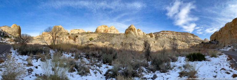 Pale sandstone spires rise behind leafless trees and snow beside a road.
