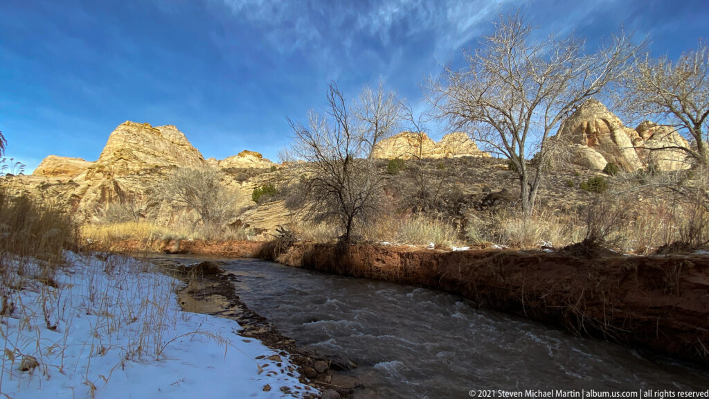 A shallow stream passes snowy banks and bare trees beneath sunlit pale sandstone cliffs.