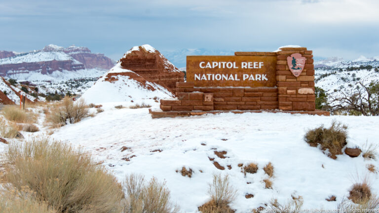 A Capitol Reef National Park sign stands on a stone base amid snow, with red cliffs behind it.
