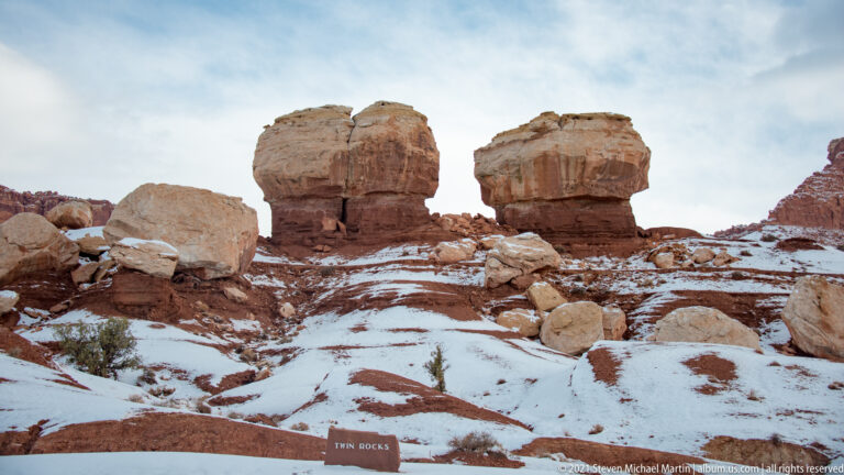 Two broad caprocks balance on narrow pedestals above a snowy slope beside a Twin Rocks marker.