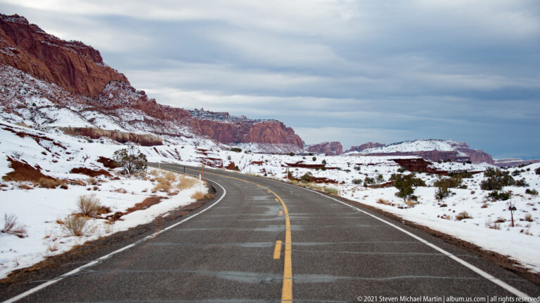 A paved road curves through snow toward red cliffs and distant mesas.