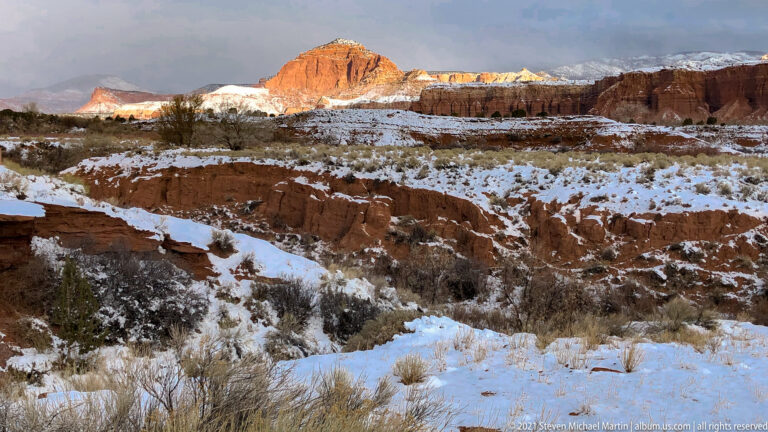 A red sandstone butte rises above snow-covered scrub and pale layered cliffs.