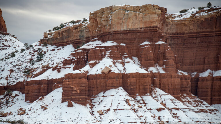 A pale caprock crowns red ribbed cliffs, with snow lodged along shelves and gullies.