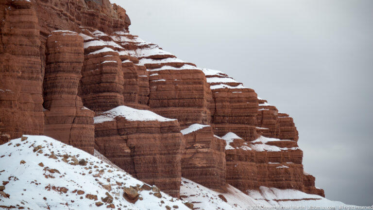 Repeated red sandstone ribs show horizontal bands and narrow snow-filled ledges across the close-up.