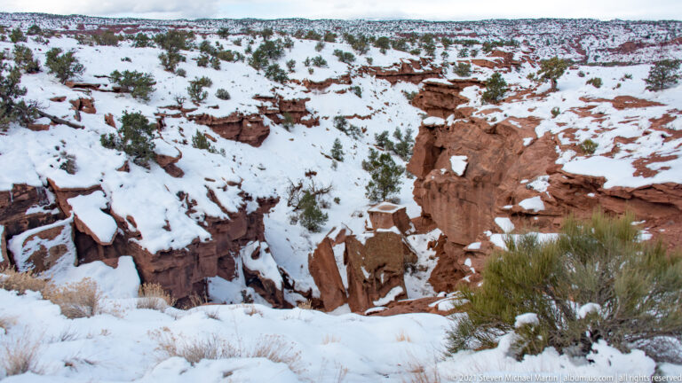 A narrow ravine cuts through snowy ground, exposing broken red rock among shrubs and grasses.