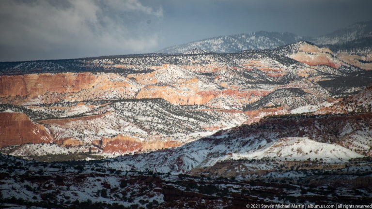 Snowy ridges and orange-red strata recede beneath dark clouds and shadow in the distance.