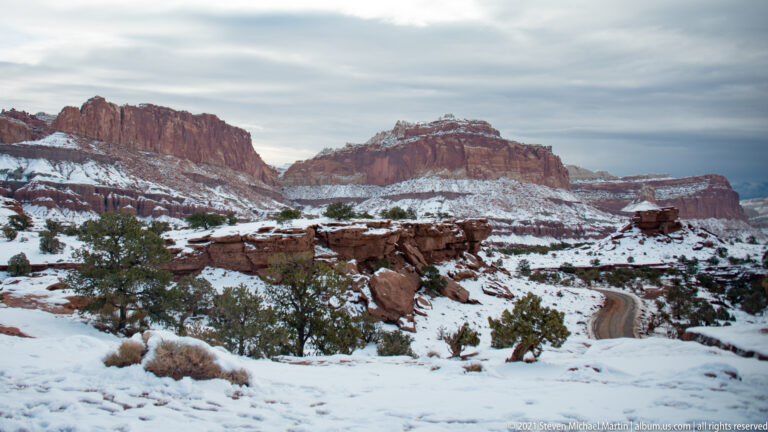 A road curves beneath snow-striped red cliffs across a juniper-dotted valley in winter.
