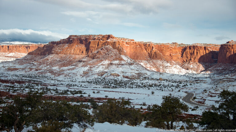 Sunlight illuminates an orange-red cliff wall above shadowed snowy ground and dark shrubs.