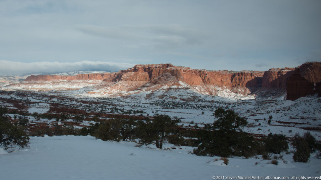 Snowy benches and junipers lead toward a broad line of red cliffs and mesas.