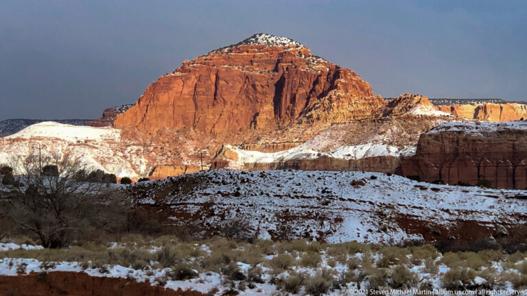 A snow-capped red sandstone butte fills the view above layered cliffs and dark shrubs.