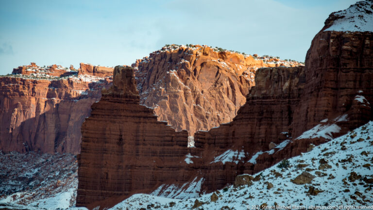 A banded sandstone tower stands between high canyon walls, with sunlit cliffs and snow behind it.