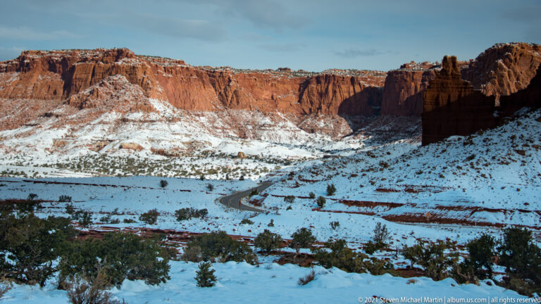 A winding road crosses a snowy valley below layered red sandstone cliffs.