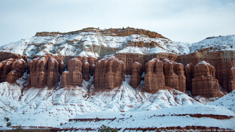 Rounded red sandstone columns rise in a row above a snowy slope, with a snow-covered ridge behind.