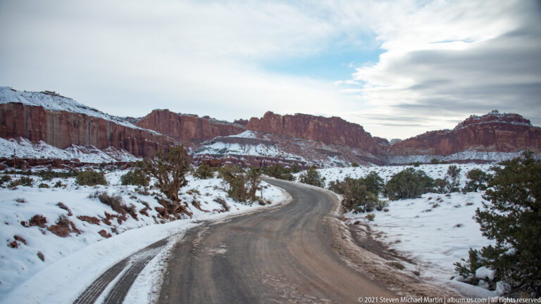 A paved road curves through snowy ground toward red sandstone cliffs and scattered juniper.