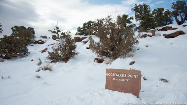 A brown stone sign in snow reads “Goosenecks Point” and “Trail 600 Feet,” with juniper behind it.
