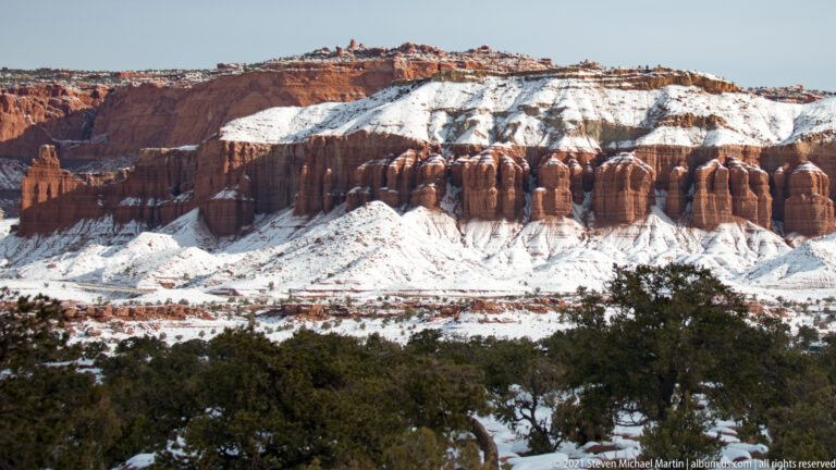Rounded red sandstone columns and a higher cliff rise above snowy ground and juniper.