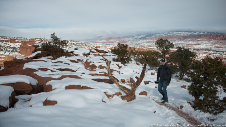 A person stands among snowy red rocks and juniper, looking toward a broad canyon.
