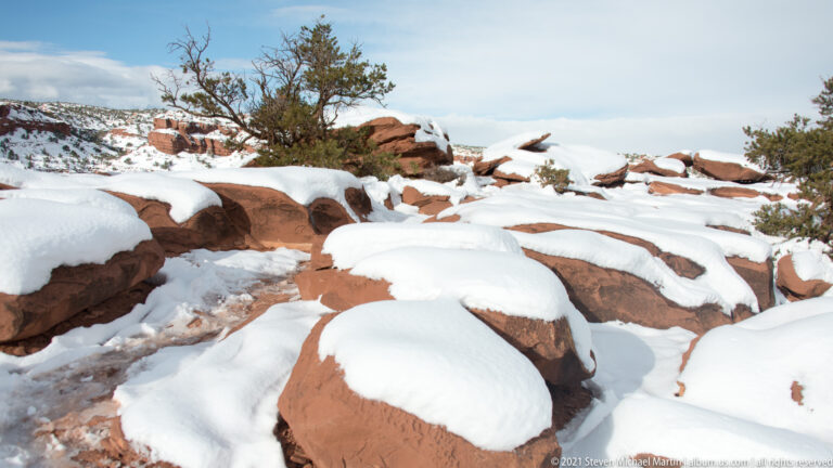 Snow fills gaps between red sandstone ledges, with a juniper and distant canyon beyond.