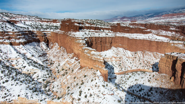A snowy canyon drops from the foreground rim toward a winding drainage and distant red plateaus.