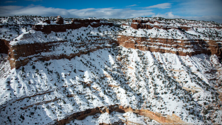 A high-angle view follows winding canyon bends through snowy red and pale rock toward distant mesas.
