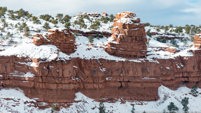 A snow-dusted red sandstone pinnacle rises above a horizontal cliff band and snowy ridge.