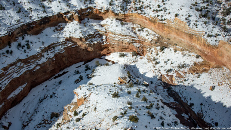A curved red canyon wall encloses a snow-covered basin seen from the rim.
