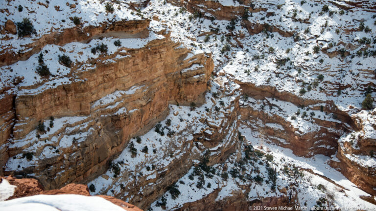 A layered red sandstone wall descends to a snowy ravine between shrub-dotted slopes.