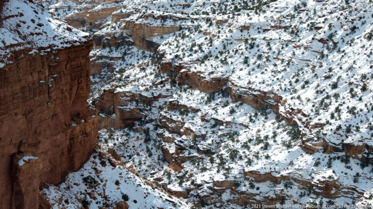 A dark cliff edge borders a narrow snow-covered channel winding through broken red rock slopes.