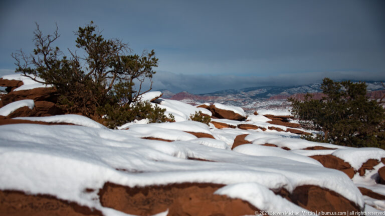 Snow-covered red rock ledges and juniper frame a hazy canyon and plateau in the distance.