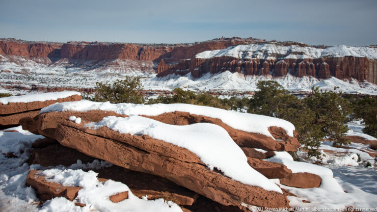 A snow-covered red sandstone ledge crosses the foreground below distant canyon walls and juniper.