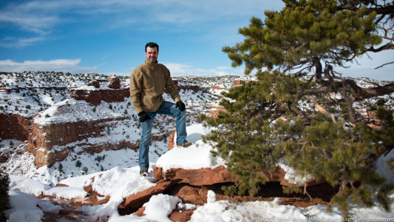 A man in a brown jacket stands on a snowy sandstone ledge above a canyon, beside a juniper.