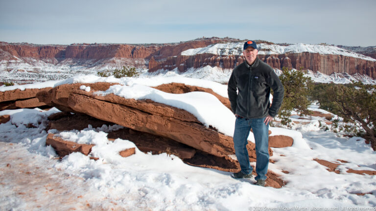 A man in a dark jacket and cap stands beside a broad snow-covered sandstone slab.