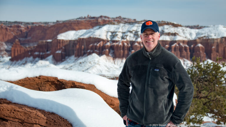 A smiling man in a dark fleece and blue cap poses before snowy red canyon walls.