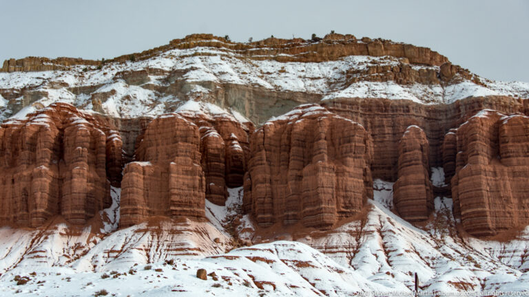 Rounded red columns rise beneath a high layered cliff, with snow tracing ledges and slopes.