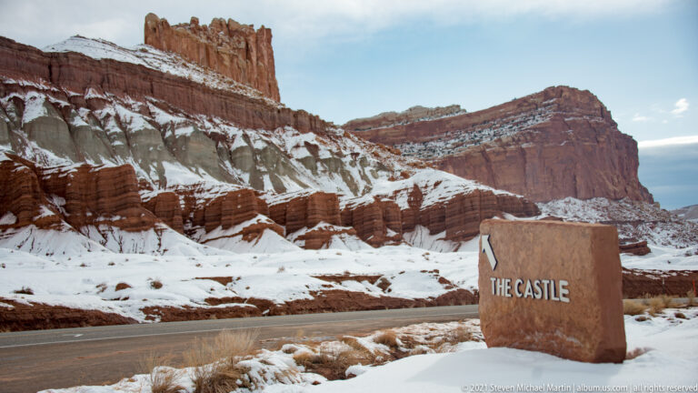 A roadside marker reading “The Castle” stands before the snow-covered red rock formation.