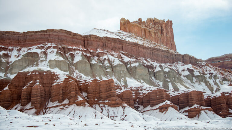 Narrow red sandstone spires rise above pale eroded slopes and rounded formations beneath a blue sky.