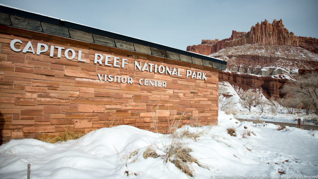 “Capitol Reef National Park Visitor Center” is lettered on a stone wall beneath red sandstone spires.