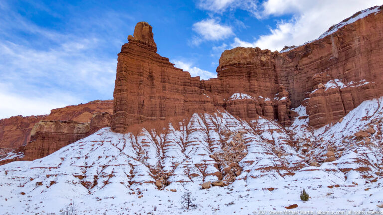 Tall banded sandstone pinnacles rise from a snowy slope beneath blue sky, beside a high cliff.