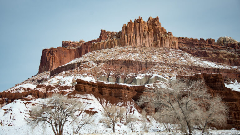 Red sandstone spires rise above snow-dusted ledges and leafless shrubs against a clear sky.